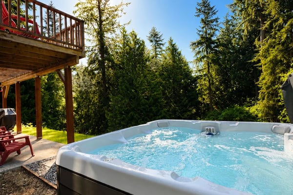 Hot tub with bubbling water on a sunny deck surrounded by tall evergreen trees and wooden house with red railing