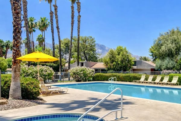 Resort pool area with spa, yellow umbrella, palm trees, lounge chairs, and mountain views under clear blue sky