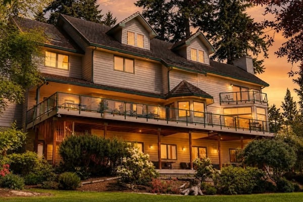 Modern farmhouse with white siding, multiple decks, and lit windows at dusk surrounded by tall trees and landscaping.