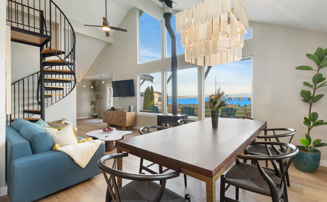 Modern open-plan living space with dining table, blue sofa, spiral staircase, and ocean views through large windows